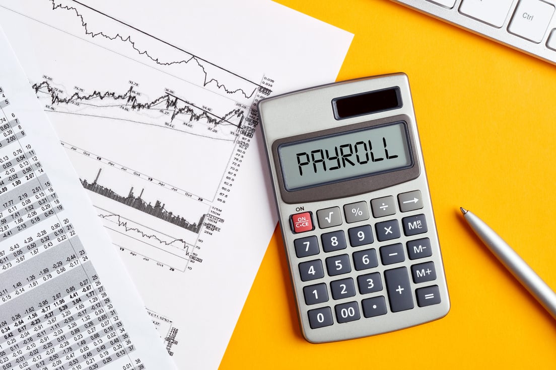 The word payroll on calculator display screen with business office desktop. Calculating or analyzing payroll expenses